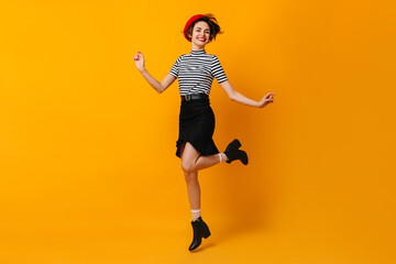 Fototapeta na wymiar Cheerful stylish french woman dancing on yellow background. Cute young lady in beret and skirt jumping with smile.