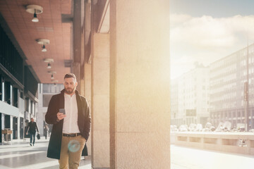 Young caucasian businesslike bearded man walking outdoor using smartphone