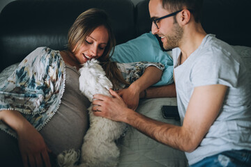 Young caucasian beautiful pregnant coule playing in bed with their dog