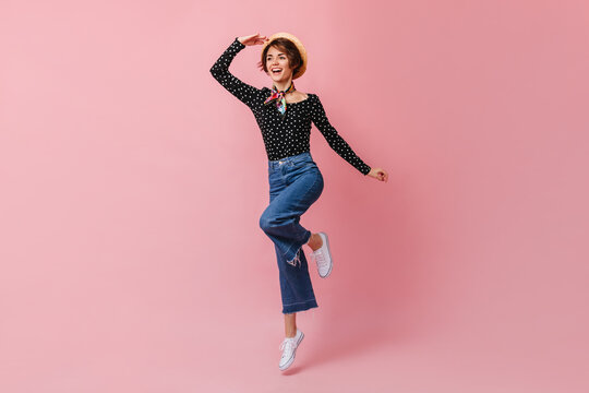 Laughing woman in straw hat looking in distance. Studio shot of amazing girl in vintage pants dancing on pink background.