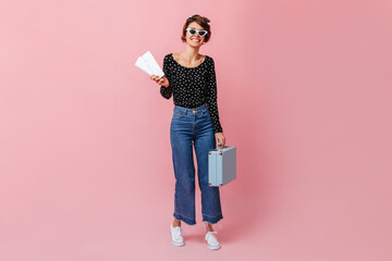 Full length view of woman in sunglasses holding tickets. Studio shot of stylish lady in jeans with blue valise.