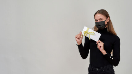 Attractive young caucasian woman wearing black outfit and protective mask looking at camera, holding gift certificate for shopping while posing isolated over gray background