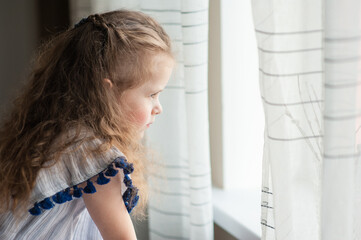 A cute little girl with long curly hair looks out the window. Portrait