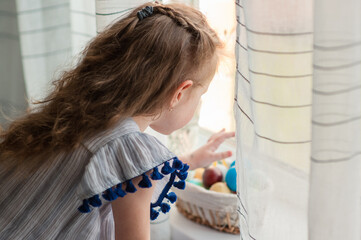 Cute little girl with long curly hair playing with colored easter eggs by the window