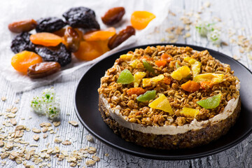 Diet cake with oatmeal and candied fruits lies on a black plate on a white wooden table next to dried fruits on paper