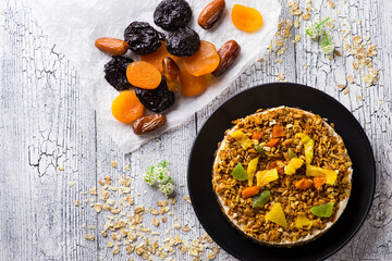 Diet cake with oatmeal and candied fruits lies on a black plate on a white wooden table next to dried fruits on paper