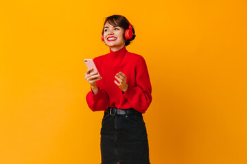 Pretty laughing woman holding smartphone on yellow background. Studio shot of good-humoured girl in headphones.