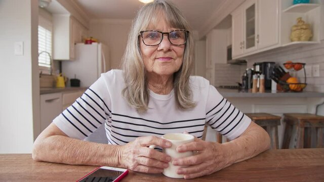 Portrait of retired Senior Caucasian woman looking at camera with her morning coffee at kitchen table. Older white woman enjoying her mug of tea at home. Slow motion dolly shot 4k
