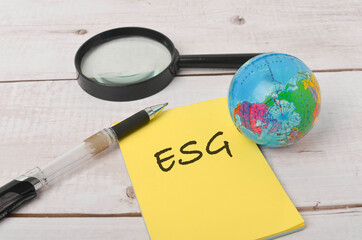 Top view of a world globe, magnifying glass and pen with written ESG on white wooden background. Selective focus.