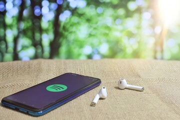 Kedah, Malaysia- 1st February 2021: Selective focus image of smartphone showing Spotify icon logo and wireless earphones. Bokeh effect background - obrazy, fototapety, plakaty