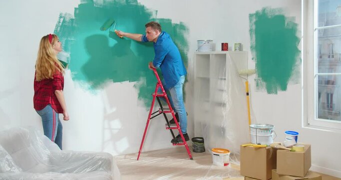 Happy Caucasian young family couple wife and husband painting wall in green color in new apartment and speaking discussing home design. Renovation, house repair and decoration concept