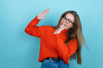 Pretty woman with long hair wearing a casual sweater and glasses over blue background scared with her arms up like something falling from above