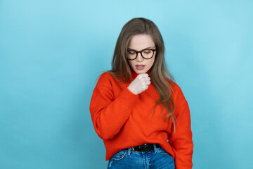 Pretty woman with long hair wearing a casual sweater and glasses over blue background with her hand to her mouth because she's coughing