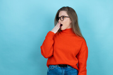 Pretty woman with long hair wearing a casual sweater and glasses over blue background covering mouth with hand, shocked and afraid for mistake. surprised expression