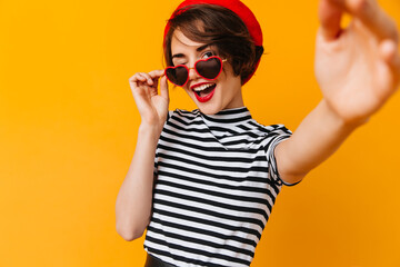Laughing girl in heart-shaped glasses taking selfie. Studio shot of french female model isolated on yellow background.
