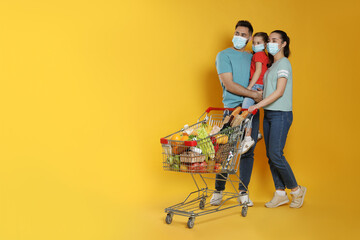 Family with protective masks and shopping cart full of groceries on yellow background. Space for...