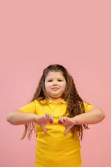 Thumbs down. Happy, smiley little caucasian girl isolated on coral pink studio background with copyspace for ad. Looks happy, cheerful. Childhood, education, human emotions, facial expression concept.