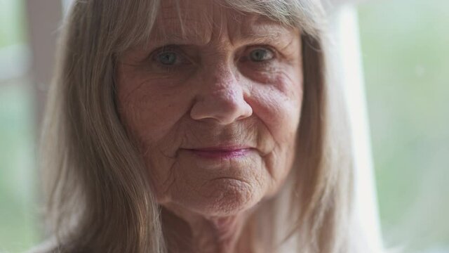 Close up portrait of friendly looking senior Caucasian woman looking at camera. Retired older white woman by a window. Slow motion 4k