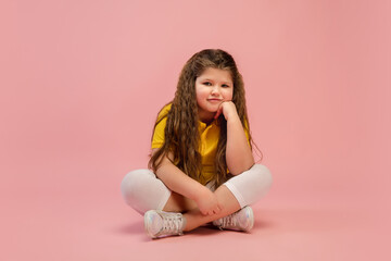 Obraz na płótnie Canvas Thinking. Happy, smiley little caucasian girl isolated on coral pink studio background with copyspace for ad. Looks happy, cheerful. Childhood, education, human emotions, facial expression concept.