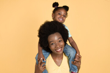 Portrait of happy African baby girl sitting on her mom's shoulders and having fun, laughing to camera on isolated yellow background