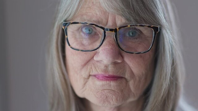 Close up portrait of sad looking senior Caucasian woman in glasses looking at camera. ECU of Retired older white woman by a window. Slow motion 4k