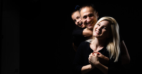 Joyful family mother, father and little girl in black clothes with dark background. Family portrait