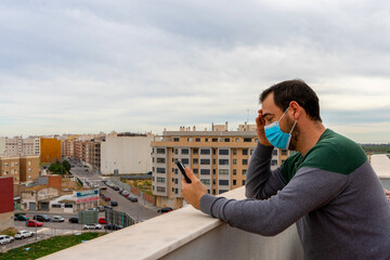 Bearded man in a gray and green sweater with a surgical mask to protect himself from the coronavirus looking at his cell phone worriedly on the terrace of his house