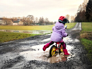Cute little toddler girl with helmet riding on run balance bike through mud and puddles. Happy...