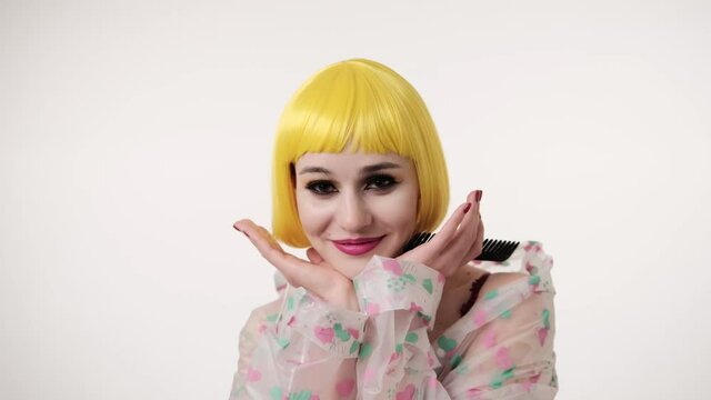 Closeup portrait of woman with colourful makeup in futuristic style smiling and wearing in yellow wig on light wall background. Creative look of woman wearing a transparent raincoat. Girl has funny wi