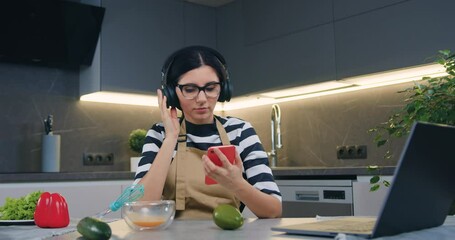 Beautiful carefree relaxed brunette in headphones choosing songs from phone playlist and whipping egg yolks in bowl with whisk in cozy kitchen,slow motion