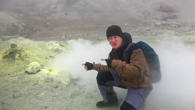 Caucasian man sits by active fumarole with sulfur surface in crater of Mutnovsky volcano on Kamchatka Peninsula, Russia. At some point, the smoke from the fumarole moves towards the person.