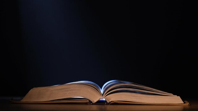 An open book lies on a table in a dark studio on a black background, a beam of light illuminates it. A stunning view of the book amid the smoke and falling snow in slow motion. Close up.