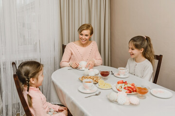 Obraz na płótnie Canvas a happy young mother and her daughters are drinking tea at the table in the kitchen of the house. breakfast with children in a happy family, happy motherhood