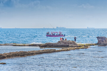 Obraz premium Pleasure boat with tourists in the sea, fishermen on the seashore. Vacation at the sea in Akko, a city on the coast of the Mediterranean Sea in Israel
