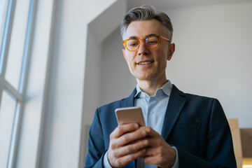 Confident mature man in stylish eyeglasses holding smartphone working online. Handsome businessman using mobile phone app, reading news, searching information standing in modern office