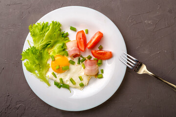 Scrambled eggs with bacon garnished with tomato and green onion on a gray concrete background near a fork.