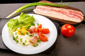 Fototapeta na wymiar Scrambled eggs with bacon, garnished with tomato and green onion and on a gray concrete background on a wooden stand next to chopped bacon and tomato.
