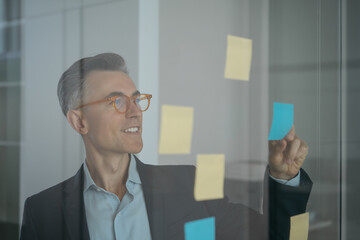 Portrait of mature scrum master wearing stylish eyeglasses using sticky notes standing near planning board in modern office. Agile methodology, scrum concept