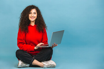 Happy young curly beautiful woman sitting on the floor with crossed legs and using laptop isolated...