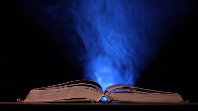 In a dark, smoky studio, a thick, open book lies on a table against a black background. Particles of dust in the light of a searchlight fly up over an open book. Slow motion. Close up.