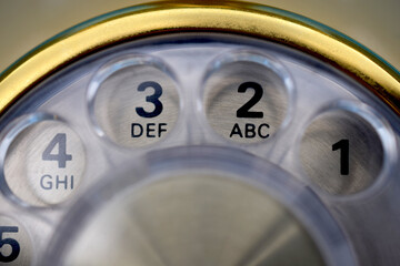 Close up view of rotary dial telephone. Numbers and letters visible. Gold and silver metal, clear plastic. For concepts related to phones, communication, old, innovation, history, and technology.