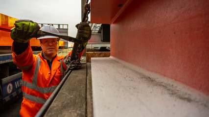 Fototapeta premium Male Driver Wearing Full PPE Chaining Down a load of Steel onto a Flatbed Trailer.
