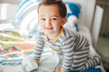 Cute adorable Caucasian kid boy sitting on bed drinking milk from kids bottle. Healthy eating drinking for little children. Supplementary food for growing babies. Candid real authentic moments.