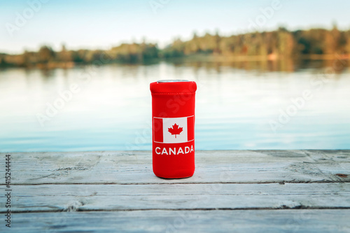 Can of beer in red cozy beer can cooler with Canadian flag standing on wooden pier by lake outdoor. Celebrating Canada Day national celebration on July 1 in nature park by the water.