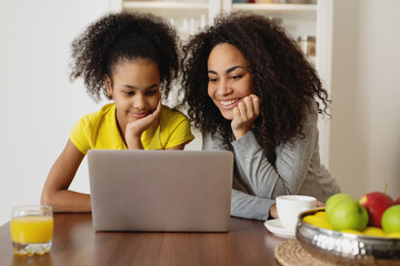 Smiling african american mother with her daughter using laptop in the kitchen.