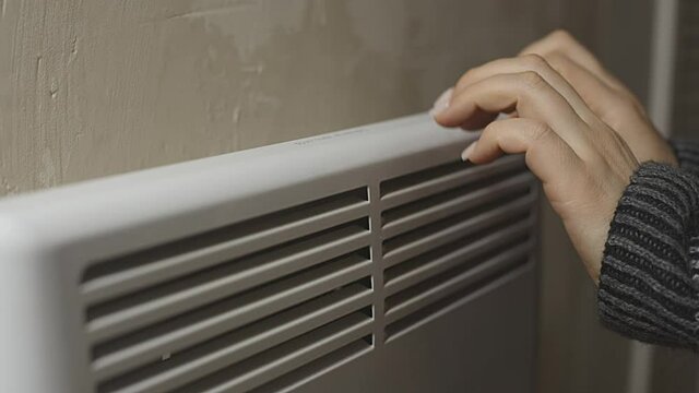 a woman warms her hands at the radiator in a cold house, problems with heating, heating the room with an electric convector