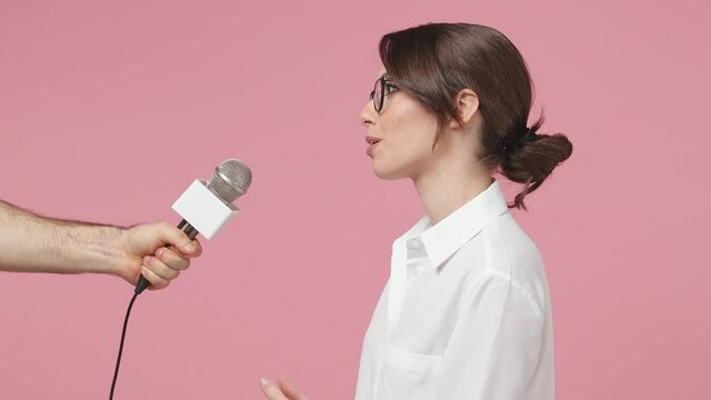Side view smiling cheerful brunette young business woman in white shirt glasses isolated on pink background studio. Achievement career wealth concept. Giving live interview in microphone to reporter