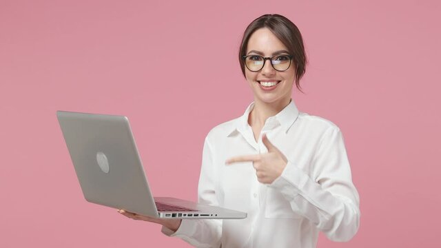 Excited young business woman 20s in white shirt glasses posing isolated on pink background studio. Achievement career wealth concept. Working point on laptop pc computer typing showing ok okay gesture