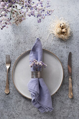 Easter violet table setting with white eggs in nest and lilac decor on grey table. Elegance dinner. Top view. Vertical format.