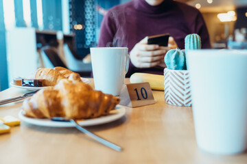 Steaming hot tea cup and fresh croissants on the table where young man using his phone during his breakfast in the hotel restaurant. Selective focus, copy space.